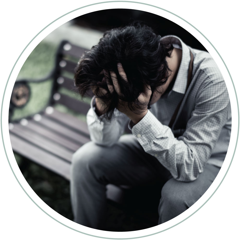 an anxious and depressed looking man at a park bench leaning forward with his hands over his head